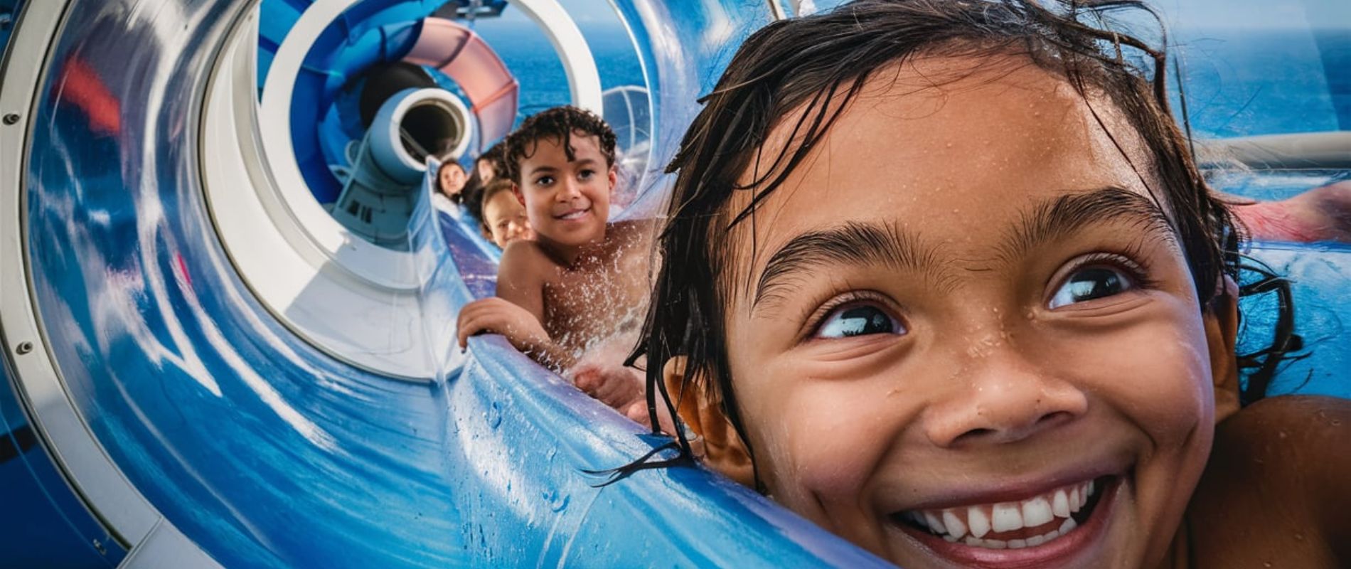 Two individuals on a water slide, experiencing the thrill of descending through a transparent tube. The slide is set against a backdrop of clear blue skies. The person on the left is wearing a black bikini and appears to be laughing with her mouth open, while the person on the right, dressed in a white top, is looking ahead with a joyful expression. Both are seated on a blue inflatable ring, which is surrounded by splashing water, indicating the slide's descent.