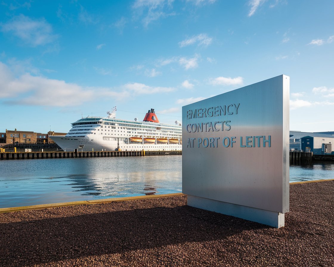 Modern Sign “Emergency Contacts at Port of Leith” with a cruise ship in the background on a sunny day