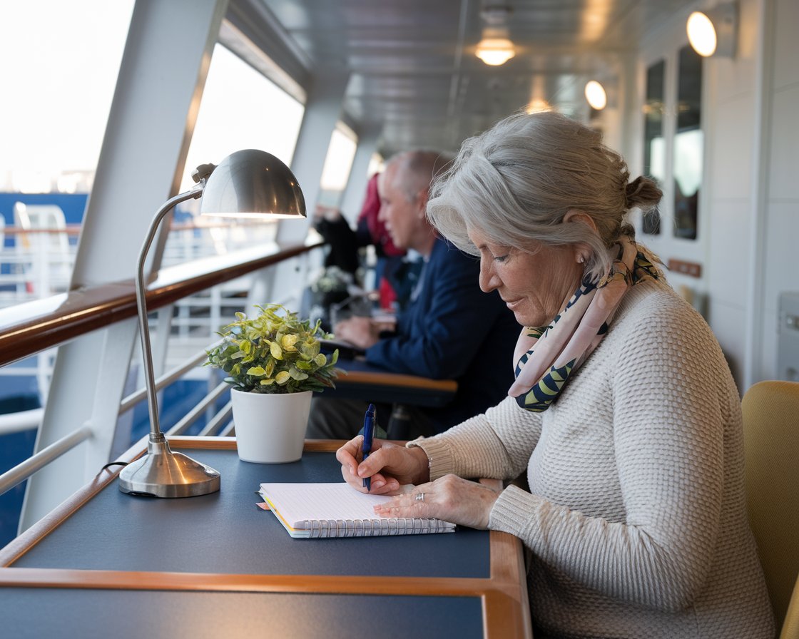 Older lady casually dressed writing at a desk on a cruise ship at Rosyth Port