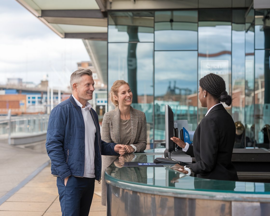 mid aged Man and mid aged lady dressed casually are at the front desk of a hotel talking to a staff member near Greenock Ocean Terminal Port