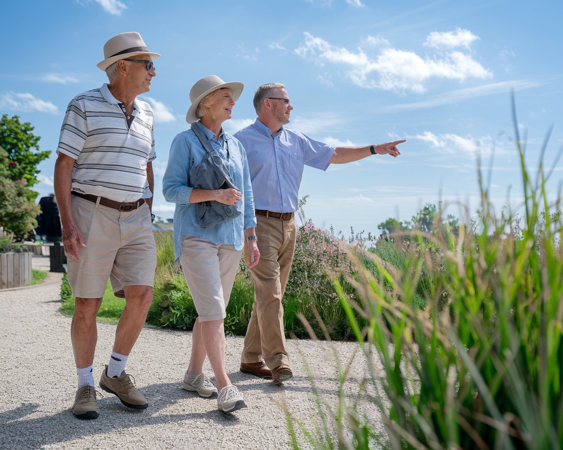 An older man and lady on a tour in Southampton. They are dressed in walking shoes and shorts with hat and sunglasses and have a tour guide pointing out the way on a sunny day.