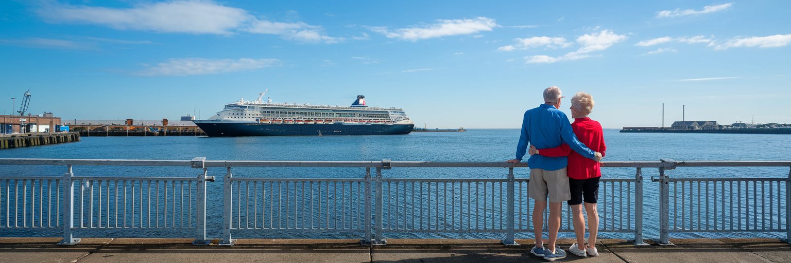 Older couple casually dressed in shorts in Port of Hull on a sunny day with a cruise ship in the background