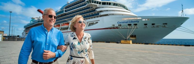 older couple casually dressed in shorts walking in Tarragona Cruise Ship Port on a sunny day with a cruise ship in the background.