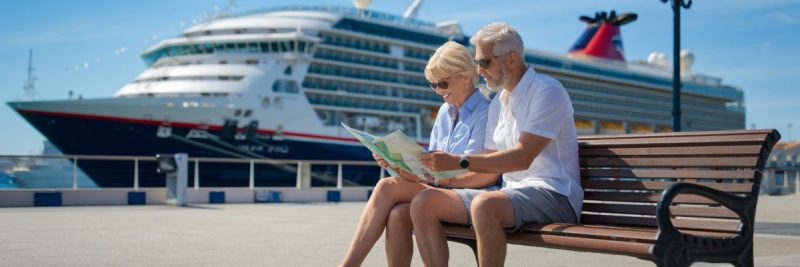 older couple sitting down, reading a map, casually dressed in shorts on a sunny day with a cruise ship in the background at Málaga Cruise Ship Port