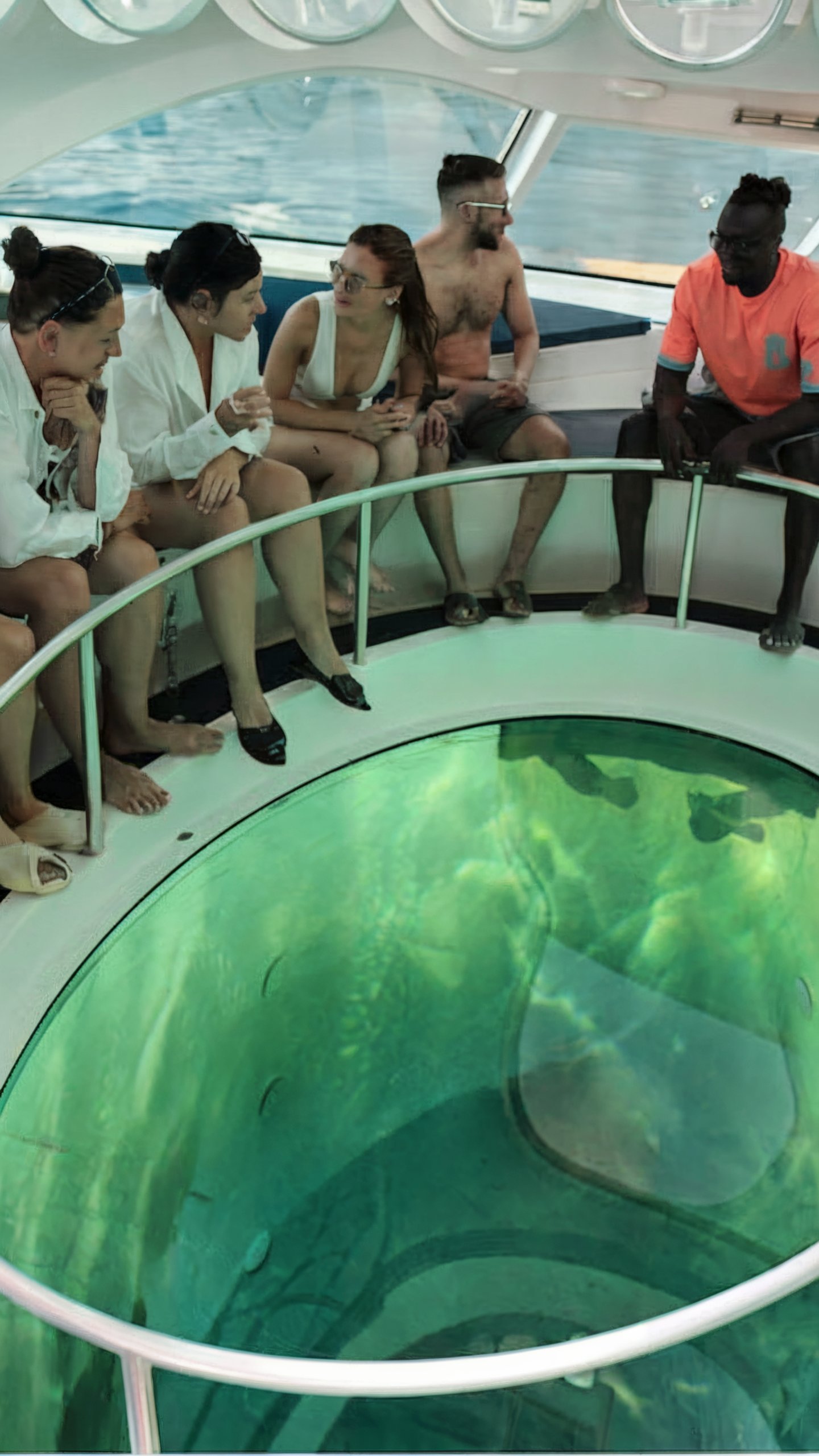 A photo of a group of people aboard a boat or yacht. They are seated around a circular glass-bottomed pool that provides a clear view of the underwater scenery. The boat's interior is modern, with white and blue tones. The group appears to be engaged in a conversation, with some individuals looking down into the pool, while others are engaged in conversation with each other. The boat's ceiling has circular lights, and the windows offer a view of the open sea., photo