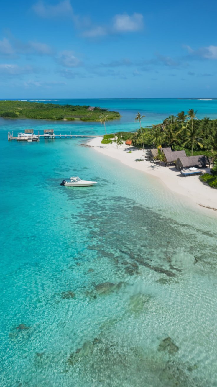 A pristine tropical beach with clear turquoise waters. On the left, there's a sandy shore with a few beach huts and a pier extending into the water. A boat is anchored near the pier. The center of the image features a boat moored at a dock, surrounded by lush greenery and palm trees. The background reveals a clear blue sky with a few scattered clouds.