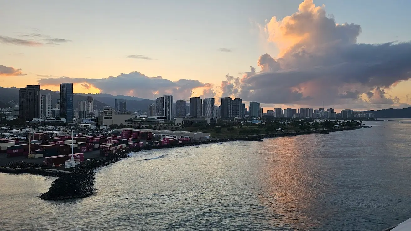 Honolulu cruise port harbor at sunrise showing downtown Waikiki skyline, container terminals, and harbor entrance as seen from arriving cruise ship on Oahu