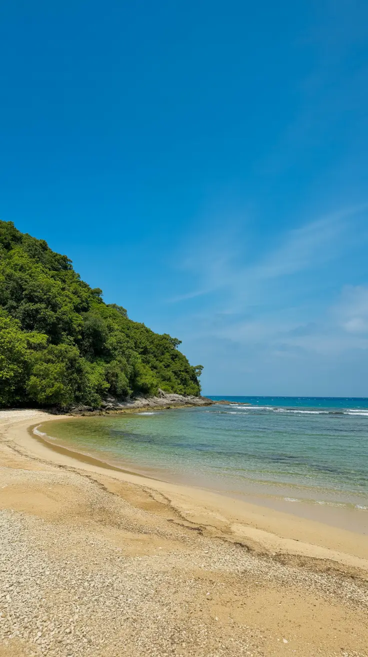 A golden, empty beach curves along turquoise water with lush green foliage under a bright blue sky.