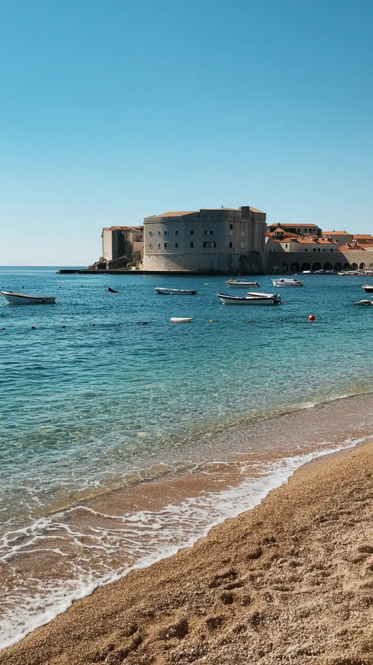 A bright, sunny day casts golden light over a small, pebbled beach near Dubrovnik's Old Town, where crystal-clear turquoise water gently laps the shore.