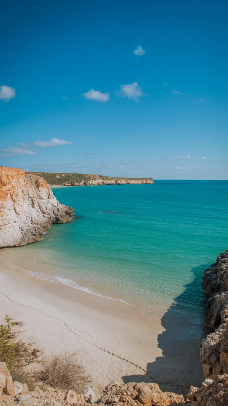 A high-end photograph captures Bahía de las Águilas from Cabo Rojo on a bright sunny day—crystal-clear turquoise water gently meets untouched white sand, with rugged cliffs framing the horizon under a vibrant blue sky.
