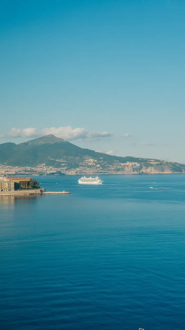A bright, sunlit photo of Naples' coastline featuring calm blue waters, a distant cruise ship, and clear skies.