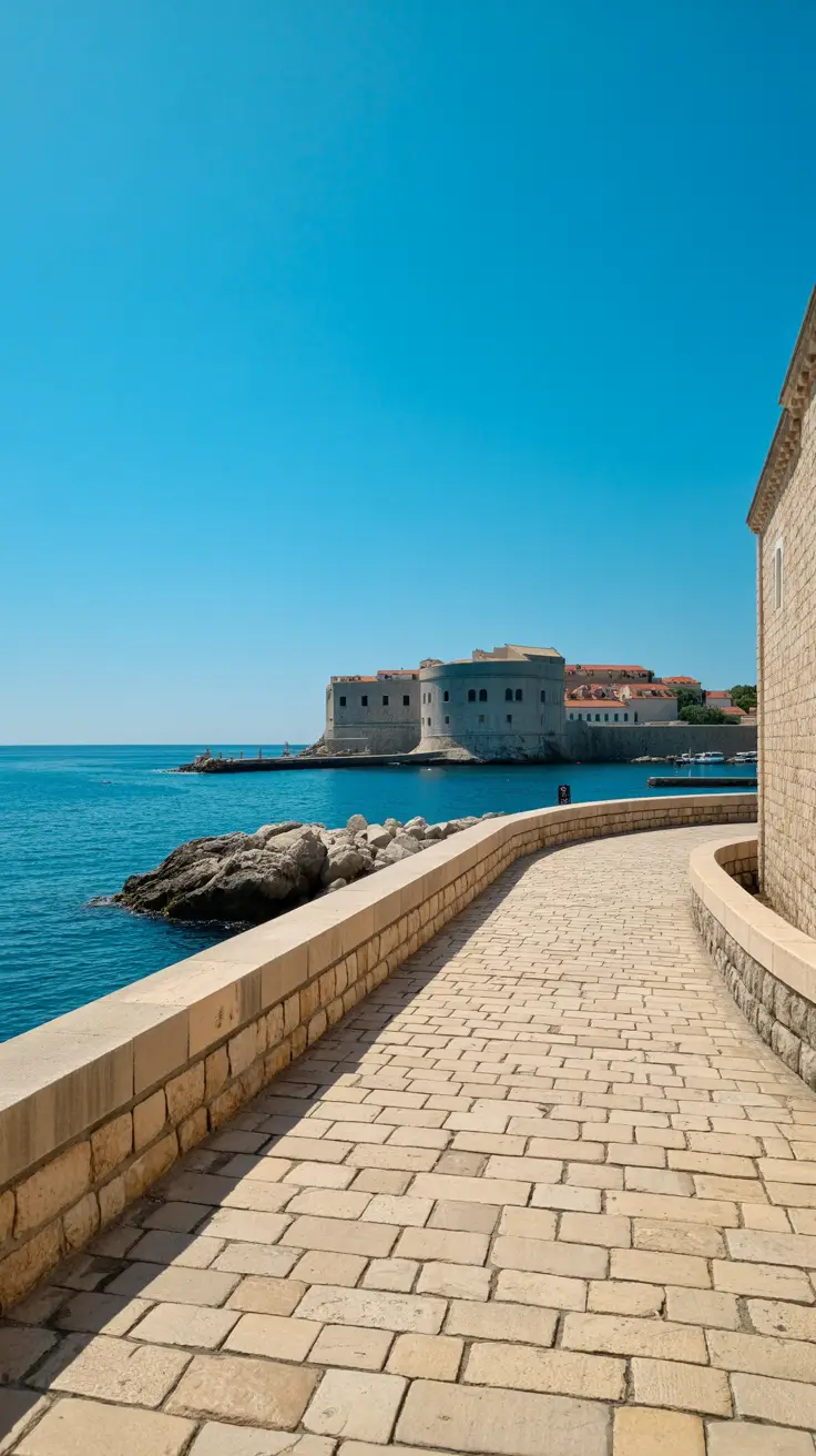 A sunlit cobblestone path winds gently along the coastline, leading from Dubrovnik Port toward the Old Town, with stone walls and vivid blue sea framing the route under a clear, bright sky.