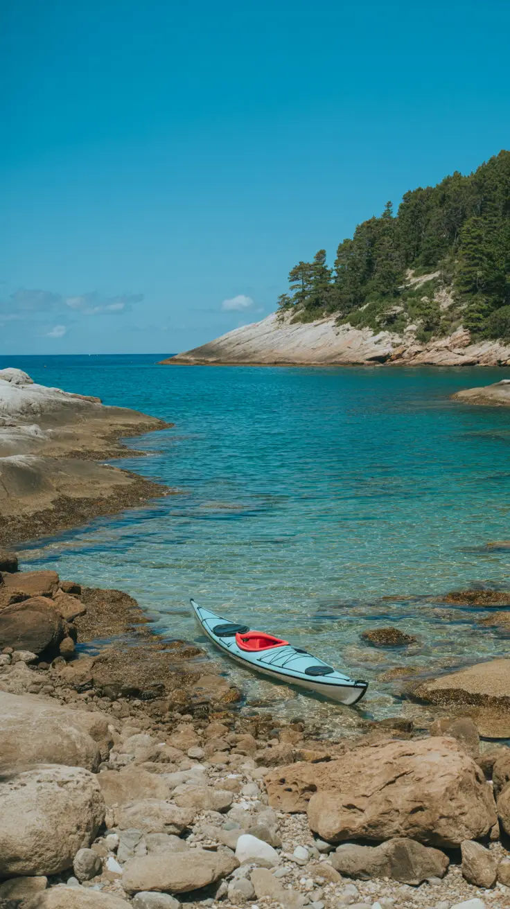 A bright, sunlit cove with crystal-clear turquoise water lapping against smooth rocks and a lone kayak resting on the shore.