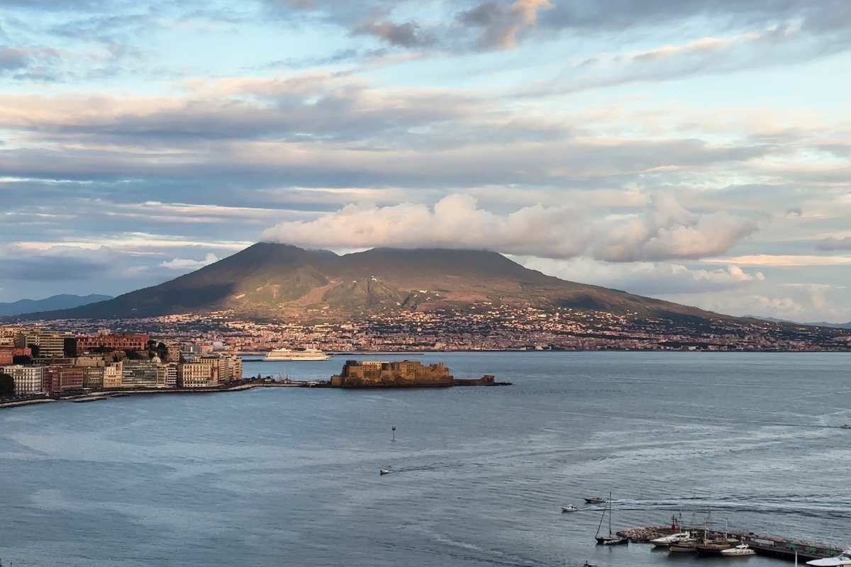 Mount Vesuvius volcano rising above Naples Bay at sunset showing both peaks with cloud formation and coastal city in foreground