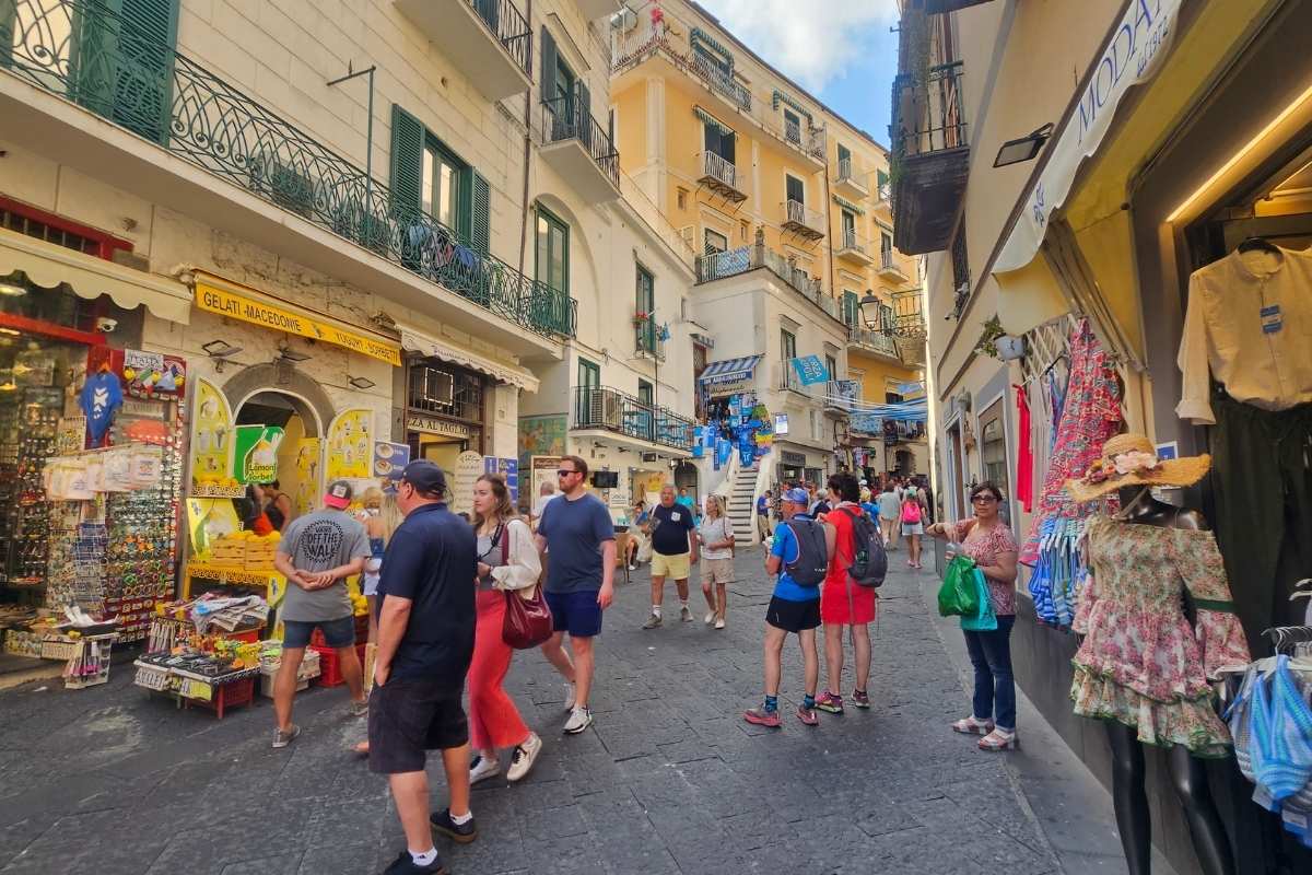 Busy pedestrian shopping street in Positano center with tourists browsing boutiques selling local ceramics, limoncello, and handmade linen clothing