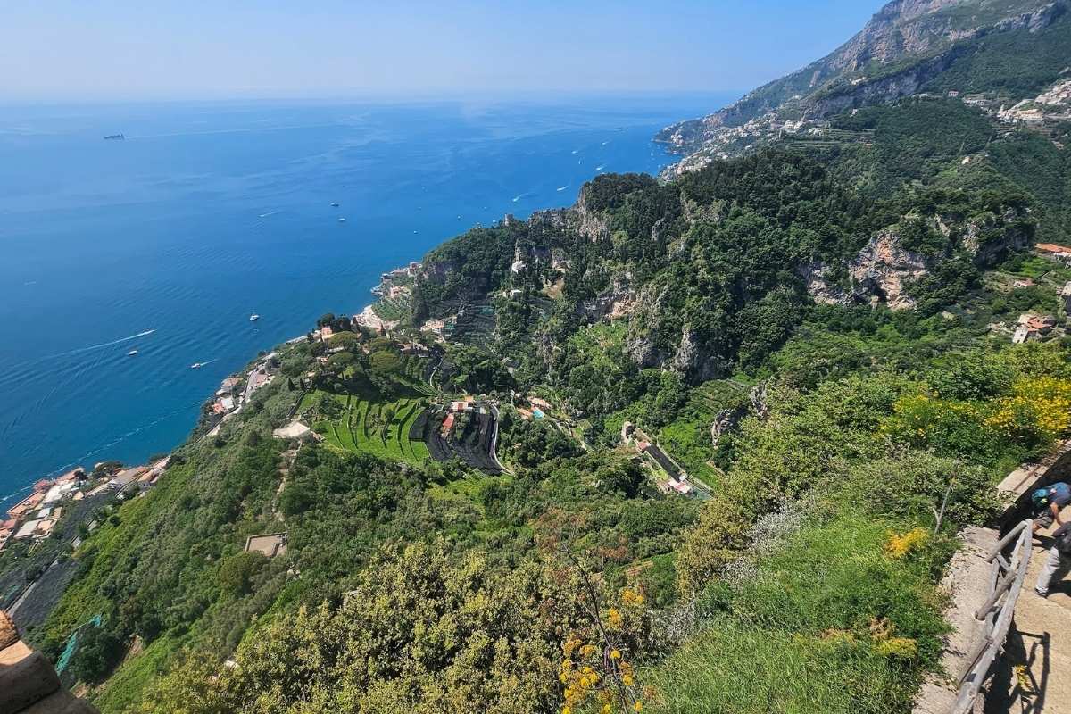 Panoramic view from Ravello gardens showing terraced vineyards, Amalfi Coast cliffs, and deep blue Tyrrhenian Sea with tourists at viewing platform