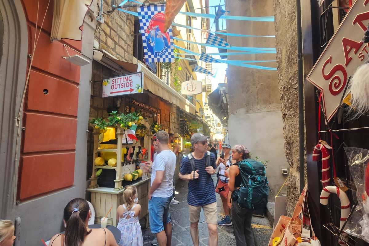 Narrow Sorrento alley with limoncello shop displaying fresh lemons, tourists sampling drinks, and colorful street decorations overhead