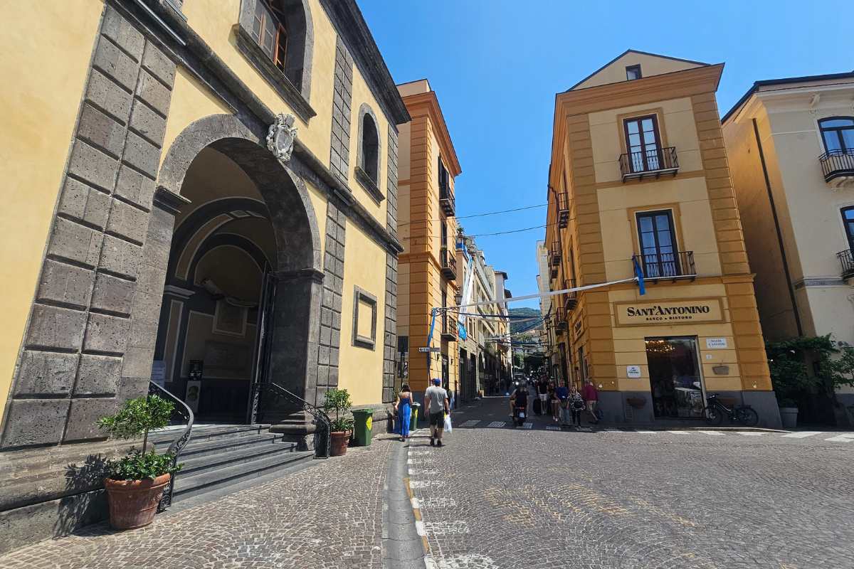 Historic Sorrento street with yellow ochre buildings, church entrance with stone steps, and tourists walking on cobblestone pavement