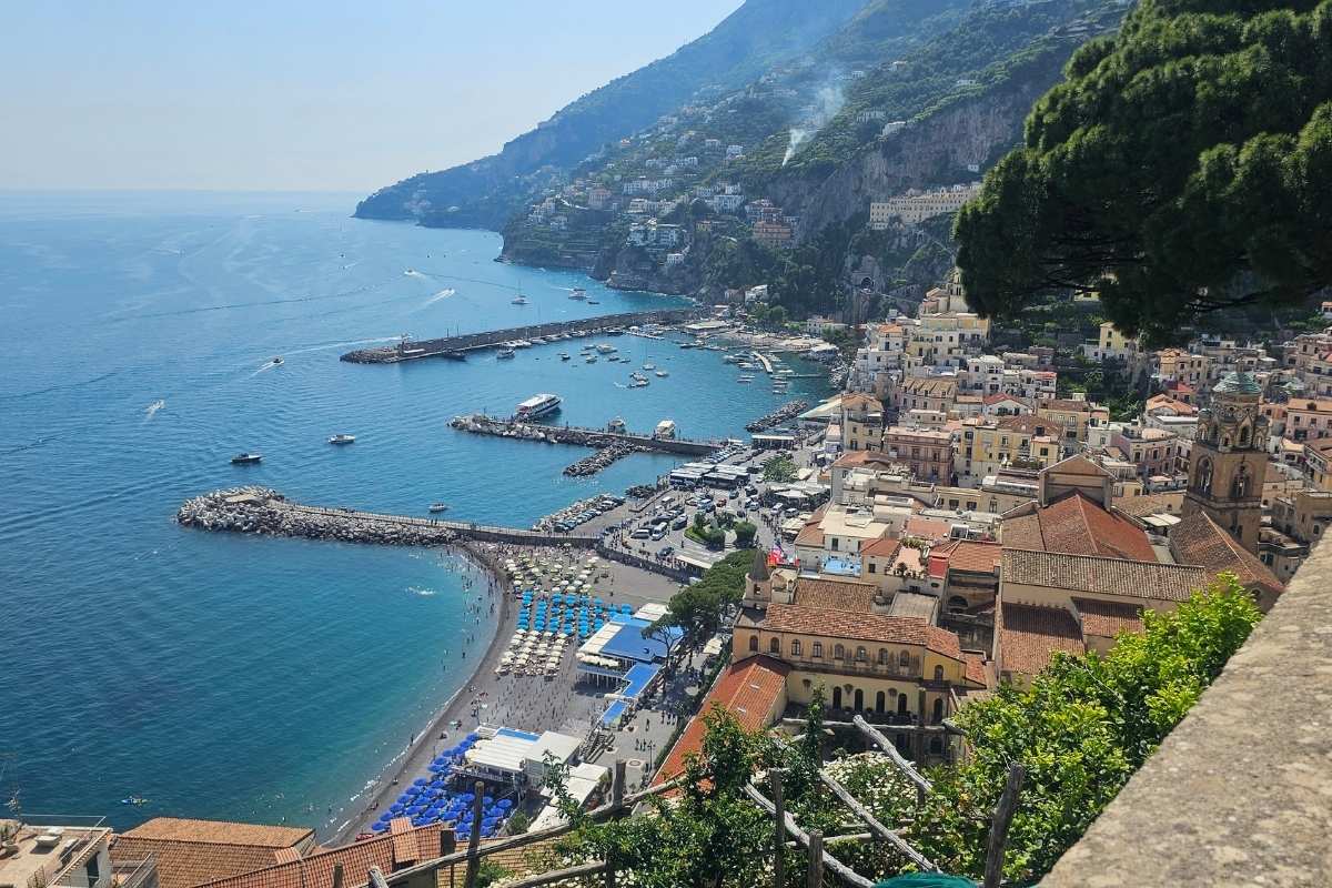 Panoramic aerial view of Positano showing Spiaggia Grande beach with blue sun loungers, boats in the harbor, and pastel-colored houses climbing the dramatic Amalfi Coast cliffs
