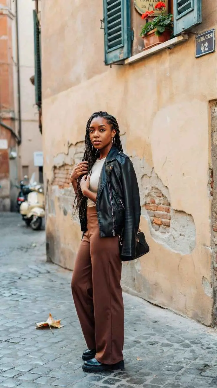 Black woman with braids in black leather jacket and brown wide leg trousers Rome cobblestone street autumn