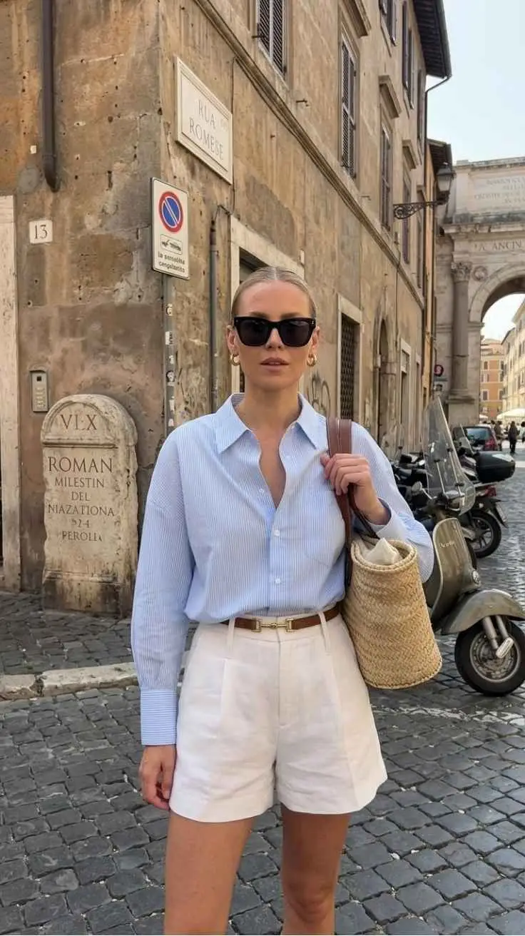 Blonde woman in blue stripe shirt and white tailored shorts with raffia bucket bag Rome cobblestones