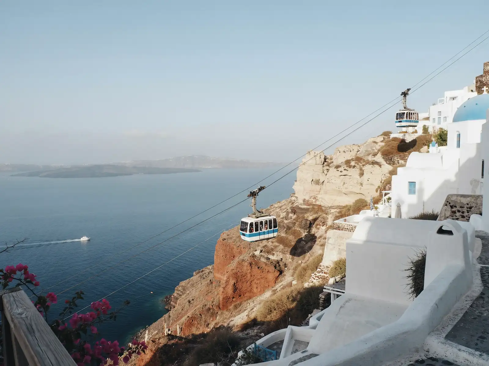 A cable car climbing the steep cliffside up to Fira, Santorini, with mules and stepped paths visible below on a sunny day.