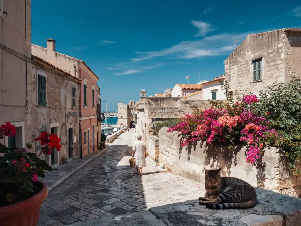Footpath leading from the port area towards the old town of Alghero, with harbour on one side and stone walls of the old town ahead, under a sunny sky.
