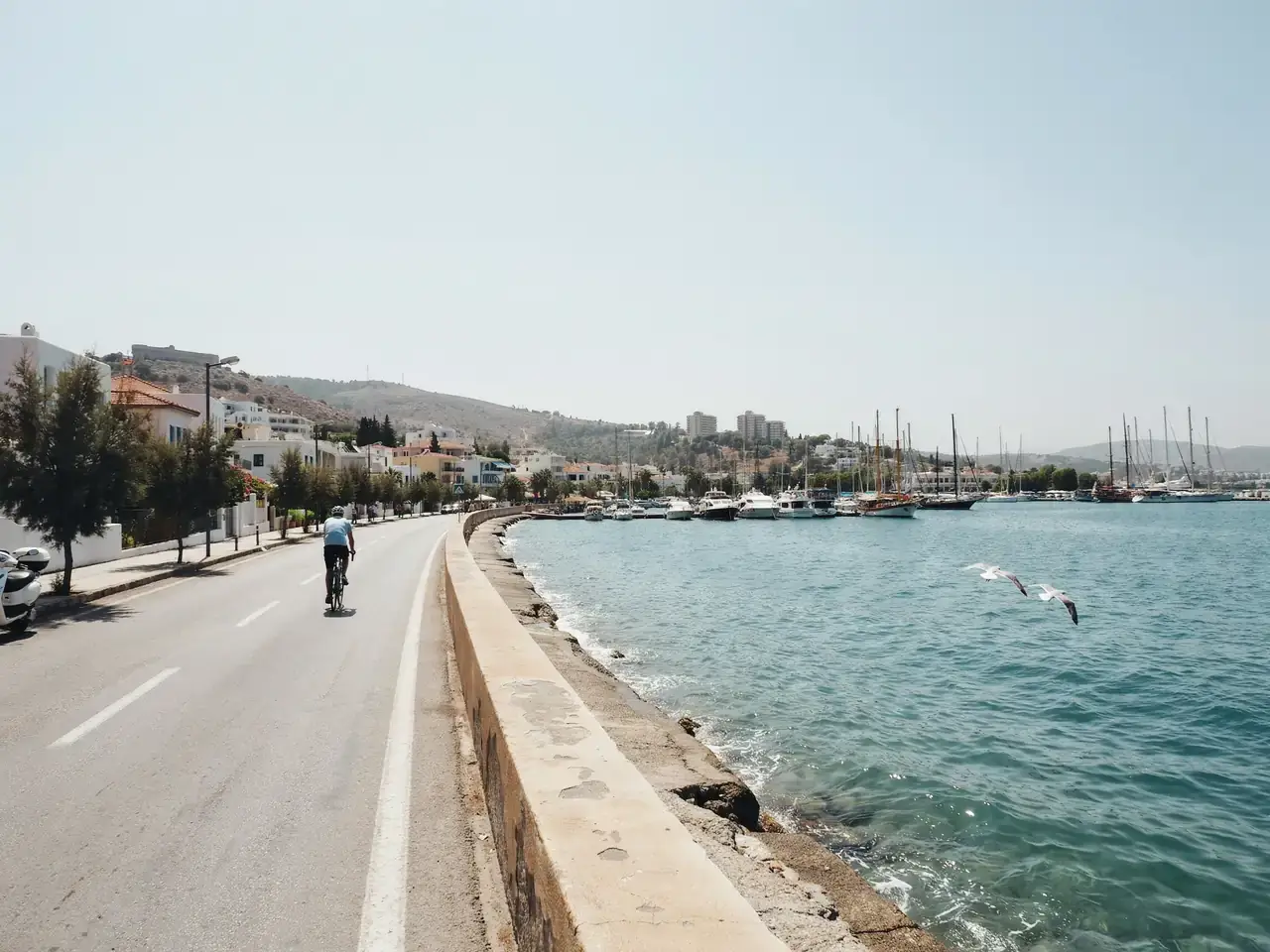 Coastal road with harbour on one side leading from Marmaris port towards the town centre, promenade and hills visible under a sunny sky
