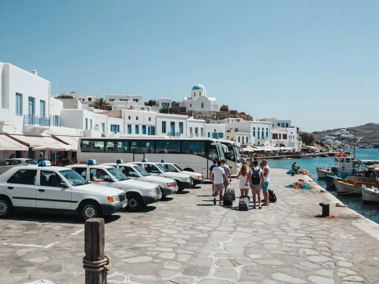 Taxis and buses lined up near Mykonos port with the town in the background on a sunny day