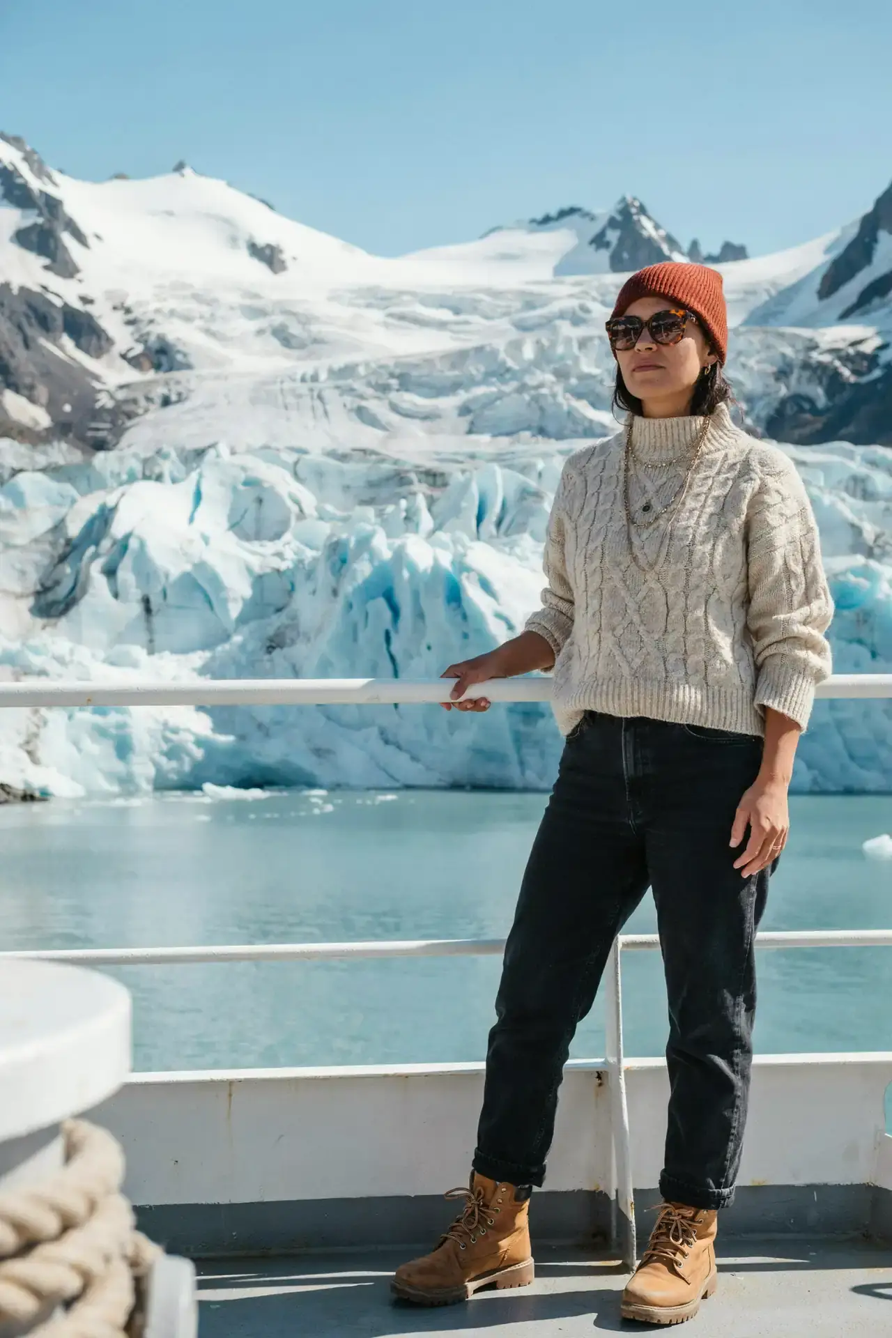 Woman in an oversized cream roll-neck jumper, dark jeans, a rust beanie and walking boots on a cruise deck in Alaska