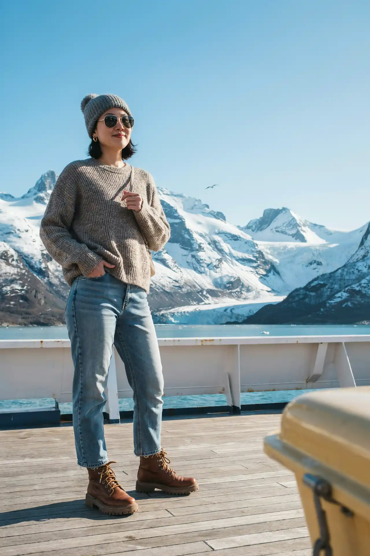 Woman in an oversized grey chunky jumper, light jeans, a woolly bobble hat and walking boots on a cruise deck in Alaska