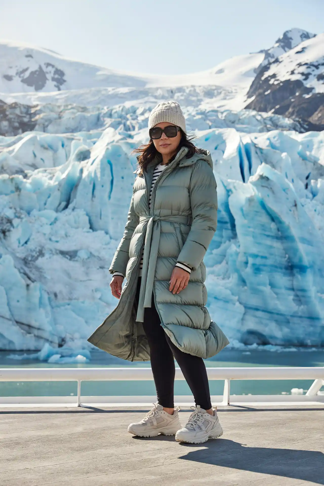 Woman in a long belted sage puffer coat over a striped top on a cruise deck in front of a glacier