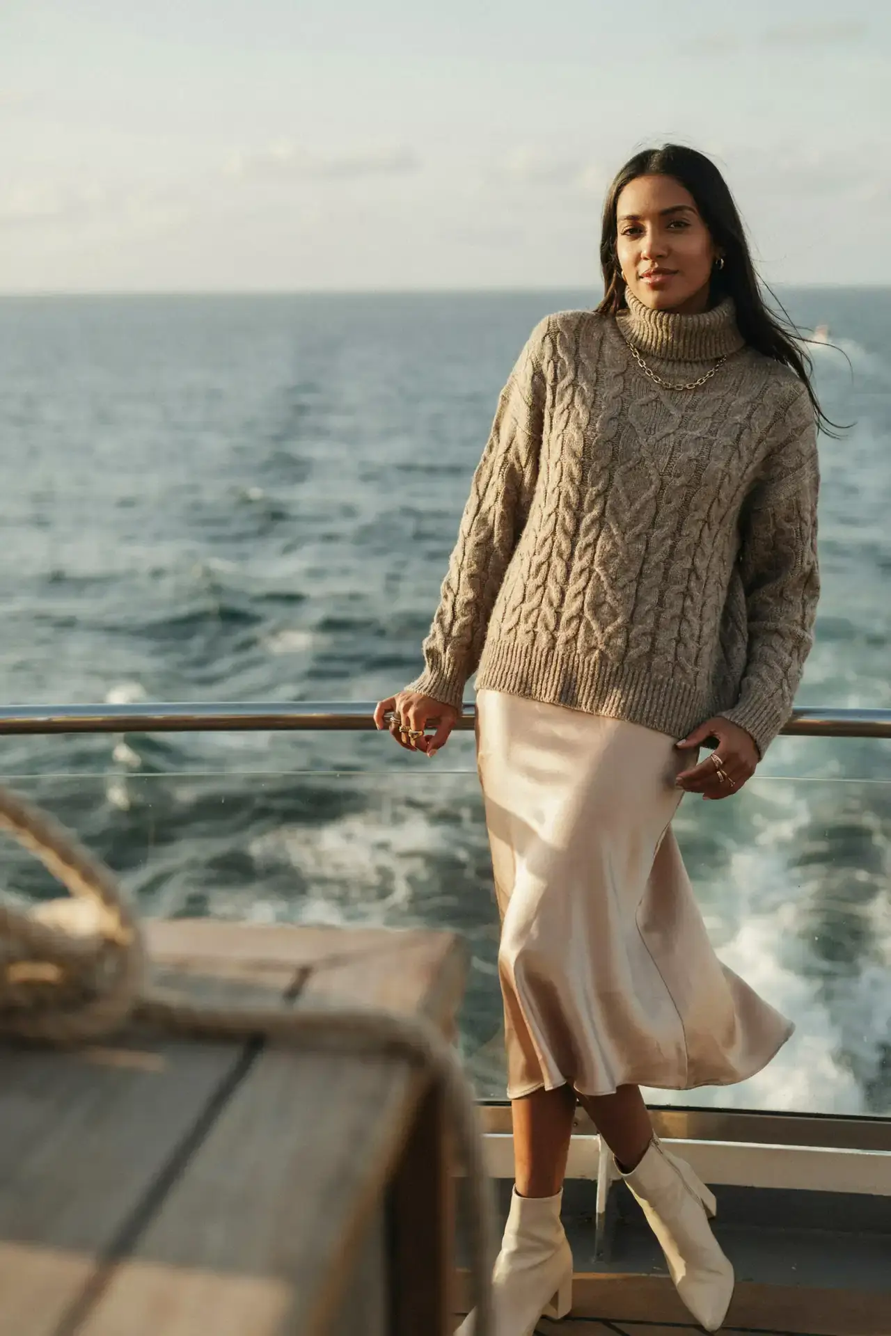 Woman in an oversized taupe turtleneck jumper, champagne satin midi skirt and cream ankle boots on a cruise deck at sunset