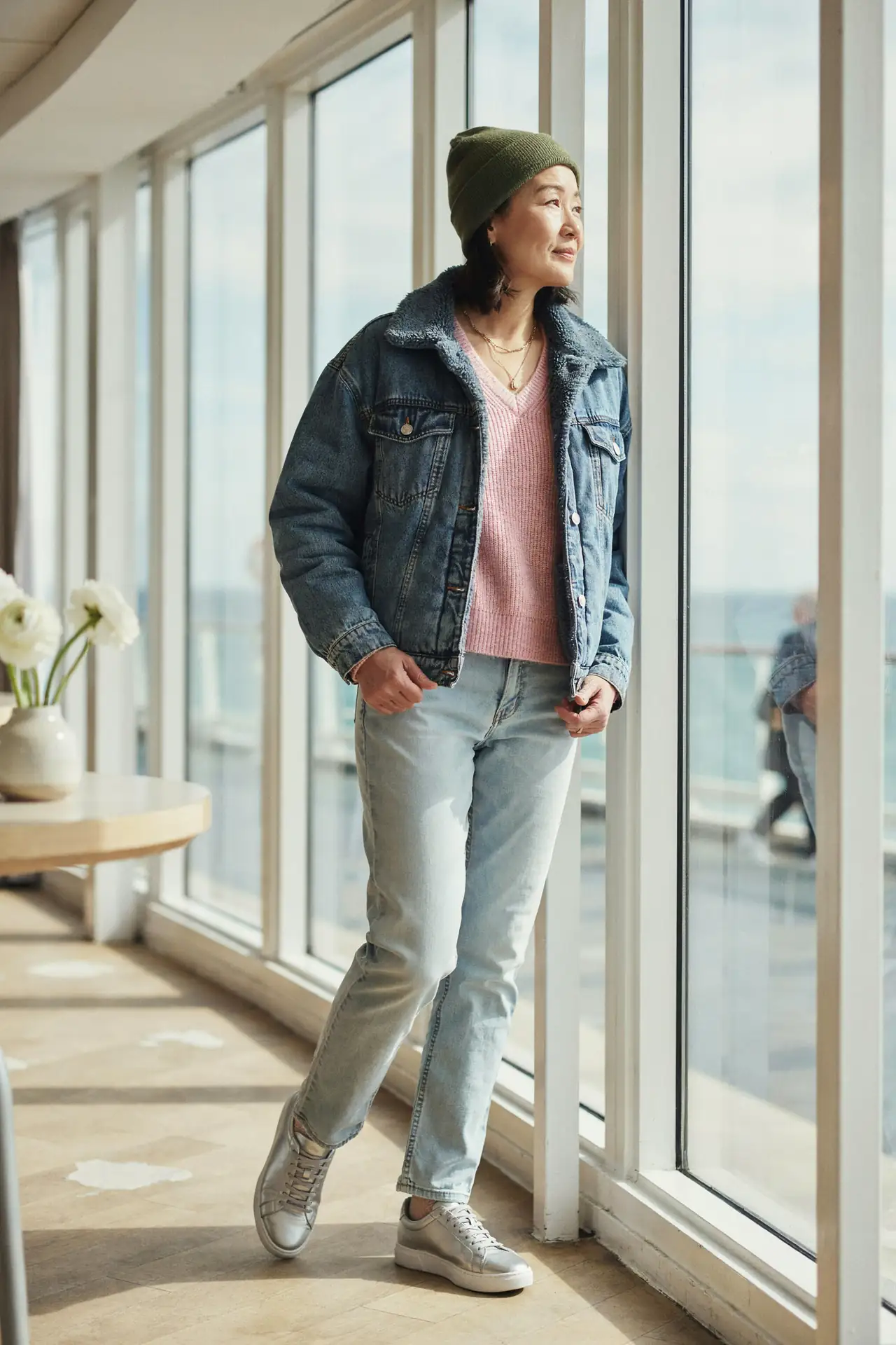 Woman in an oversized sherpa-lined denim jacket, light jeans, an olive beanie and silver metallic trainers on a cruise ship